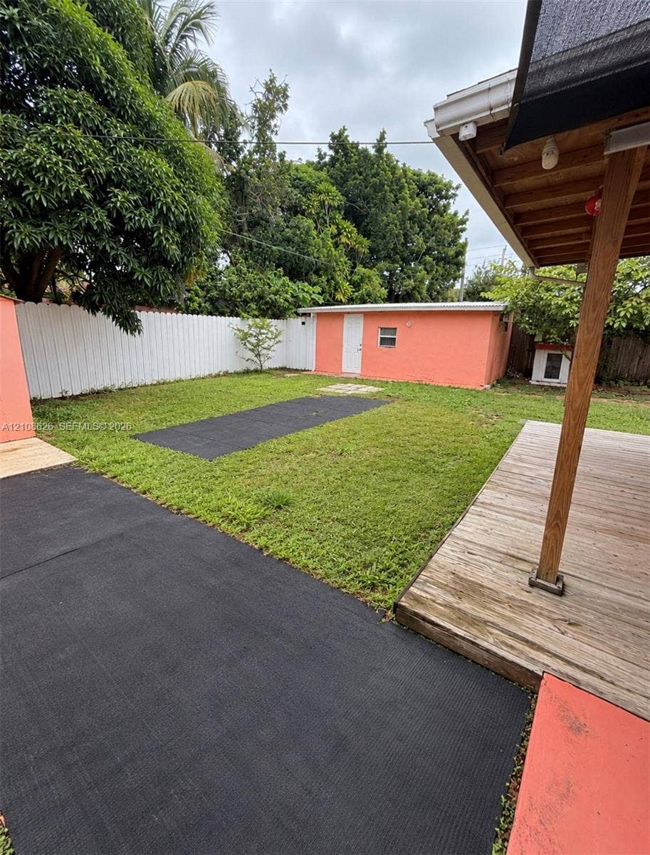 971 NE 156th Terrace, North Miami Beach, FL 33162 Backyard featuring a wood-plank deck with a covered area, a detached structure, a white privacy fence, and paved ground surfaces