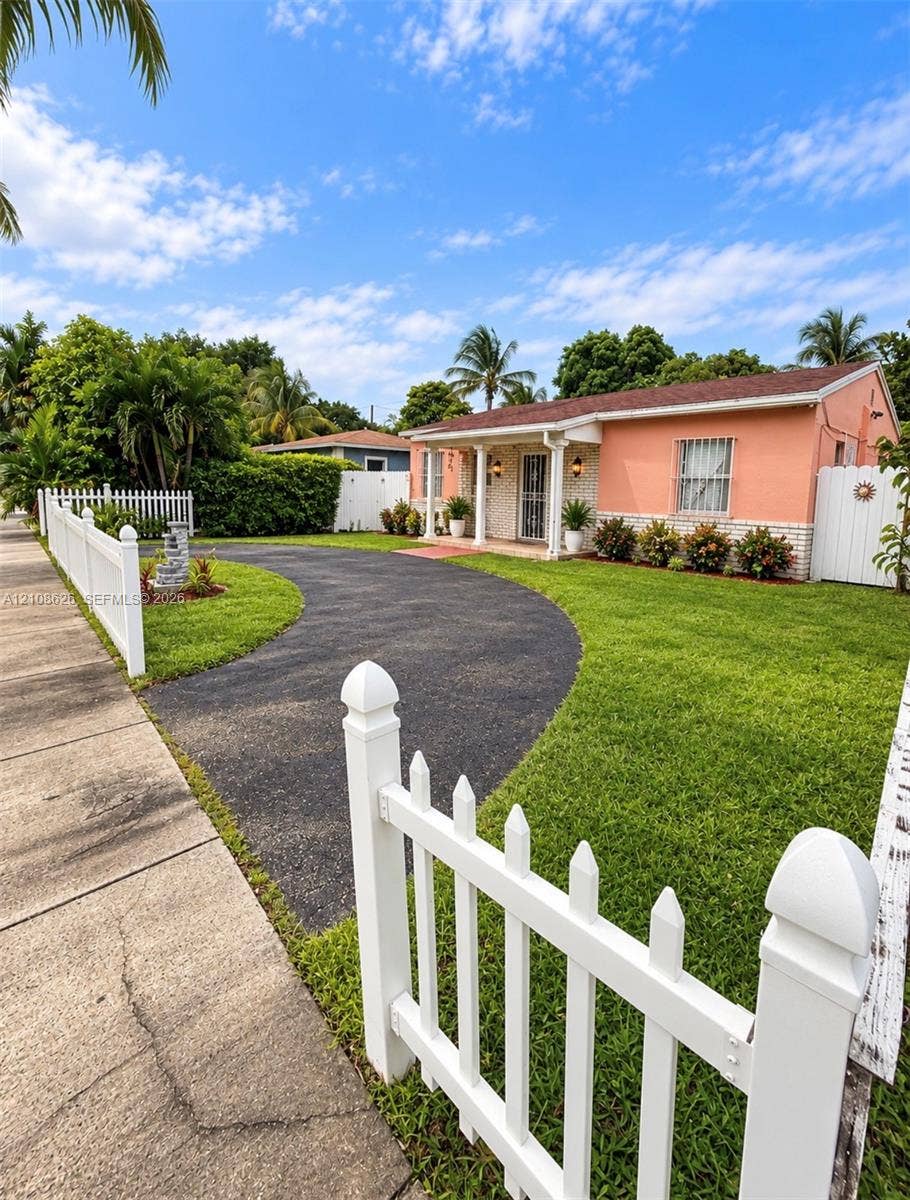 971 NE 156th Terrace, North Miami Beach, FL 33162 Single-story exterior featuring stucco and brick facade, a curved asphalt driveway, and perimeter white picket fencing