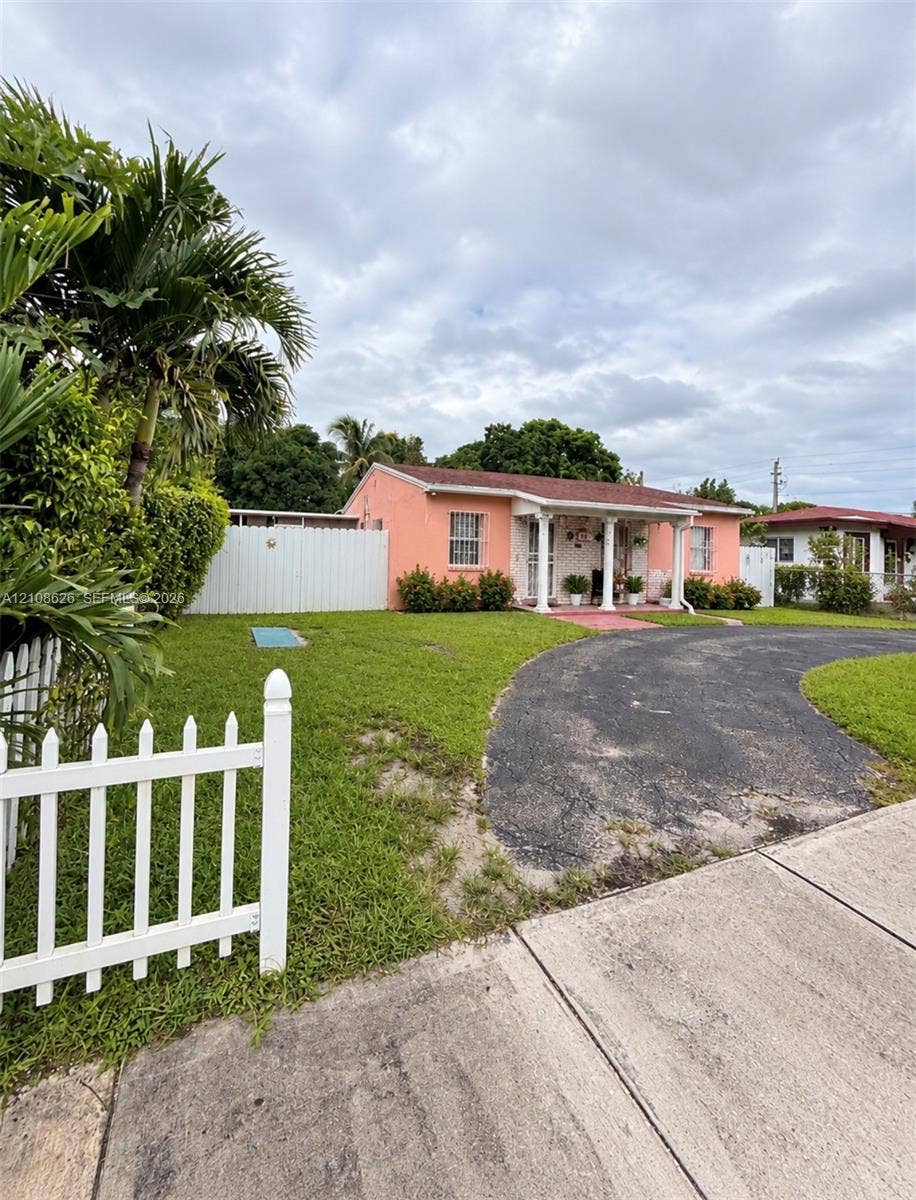 971 NE 156th Terrace, North Miami Beach, FL 33162 Stucco exterior featuring a covered front porch with decorative columns, a paved curved driveway, and white picket fencing
