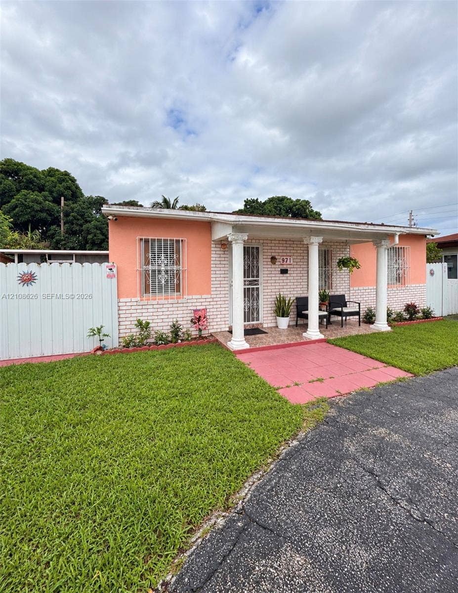 971 NE 156th Terrace, North Miami Beach, FL 33162 Exterior facade featuring brick masonry, a covered front porch with classic columns, metal window security bars, and perimeter privacy fencing
