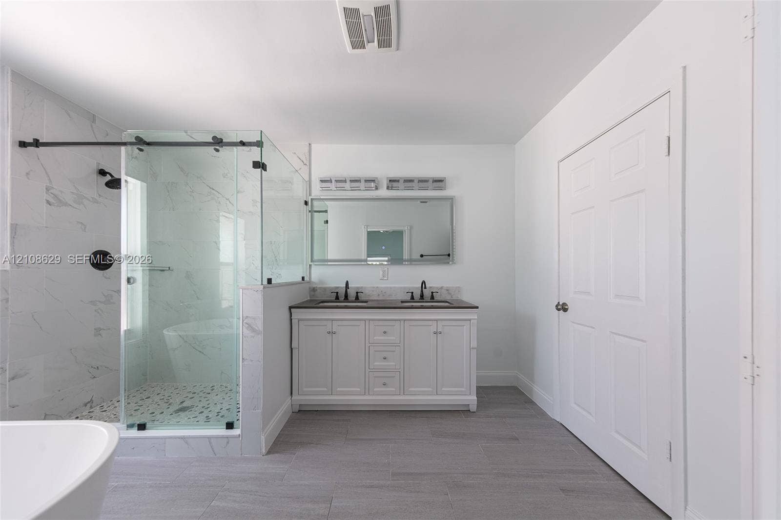 Dual-sink vanity with a solid-surface countertop, glass-enclosed walk-in shower with stone-patterned tile, freestanding soaking tub, and gray tile flooring