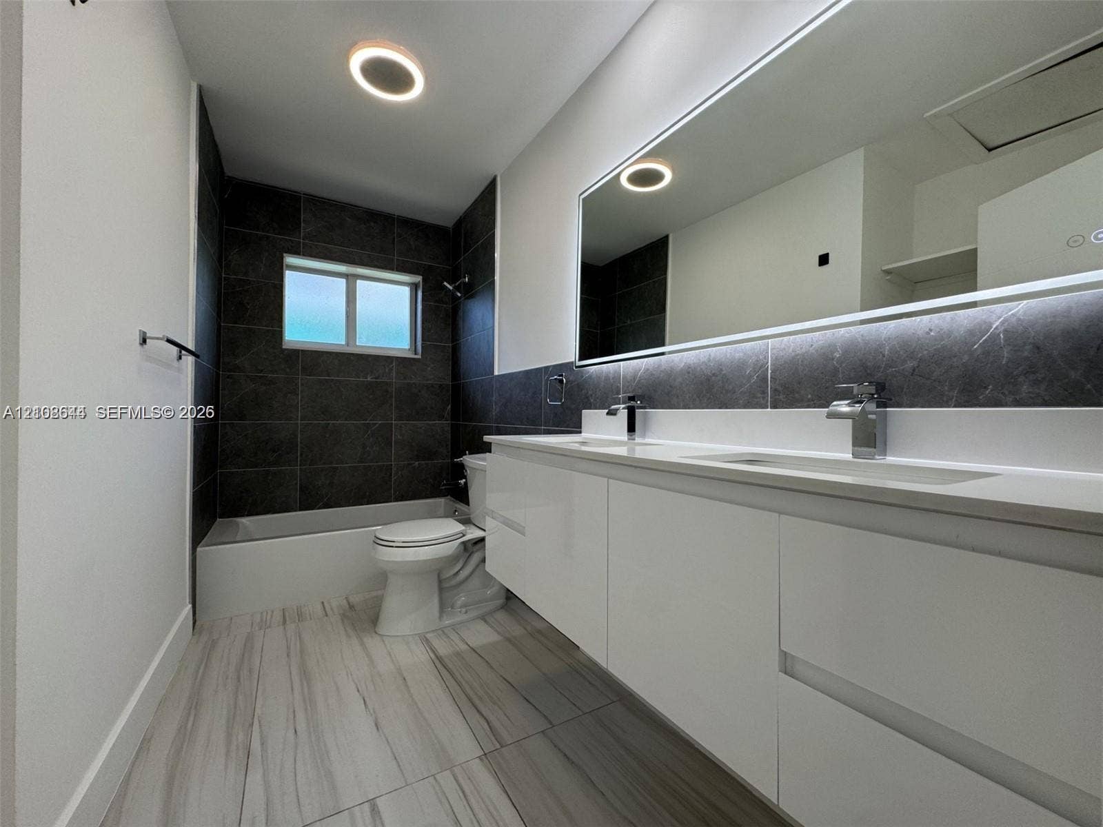 1431 N 14th Way, #rear, Hollywood, Hollywood, FL 33020 Dual-sink floating vanity with sleek white cabinetry, large illuminated mirror, contrasting dark stone-finish shower wall tile, and light-toned wood-finish flooring
