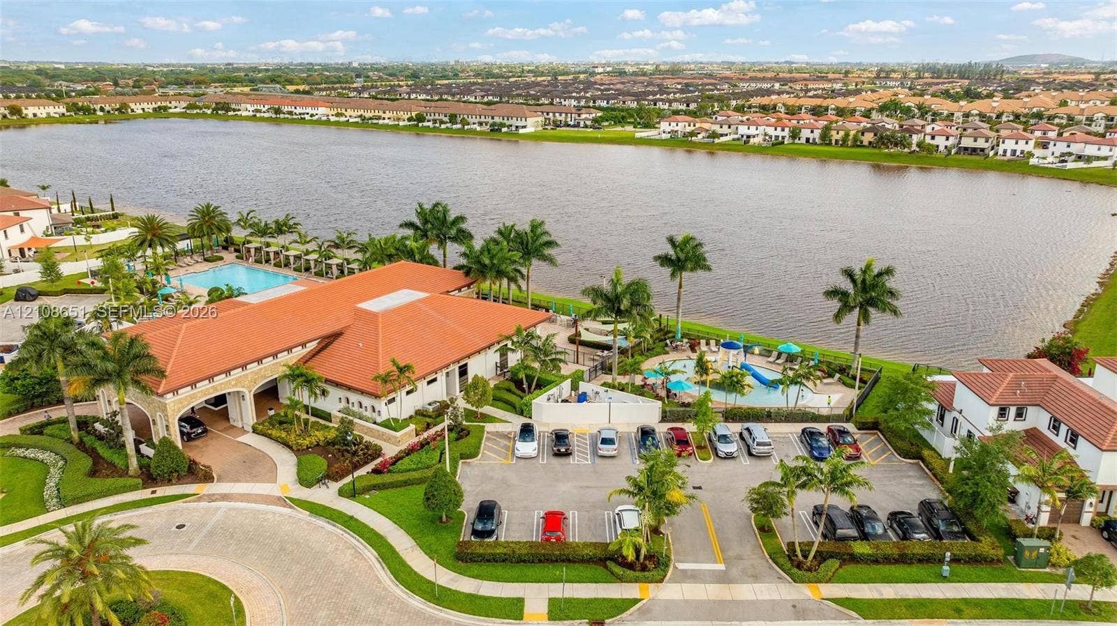 3288 W 106th Terrace, #3288, Hialeah, Hialeah, FL 33018 Waterfront clubhouse featuring a tile roof, an outdoor swimming pool, paved parking, and manicured landscaping with palm trees