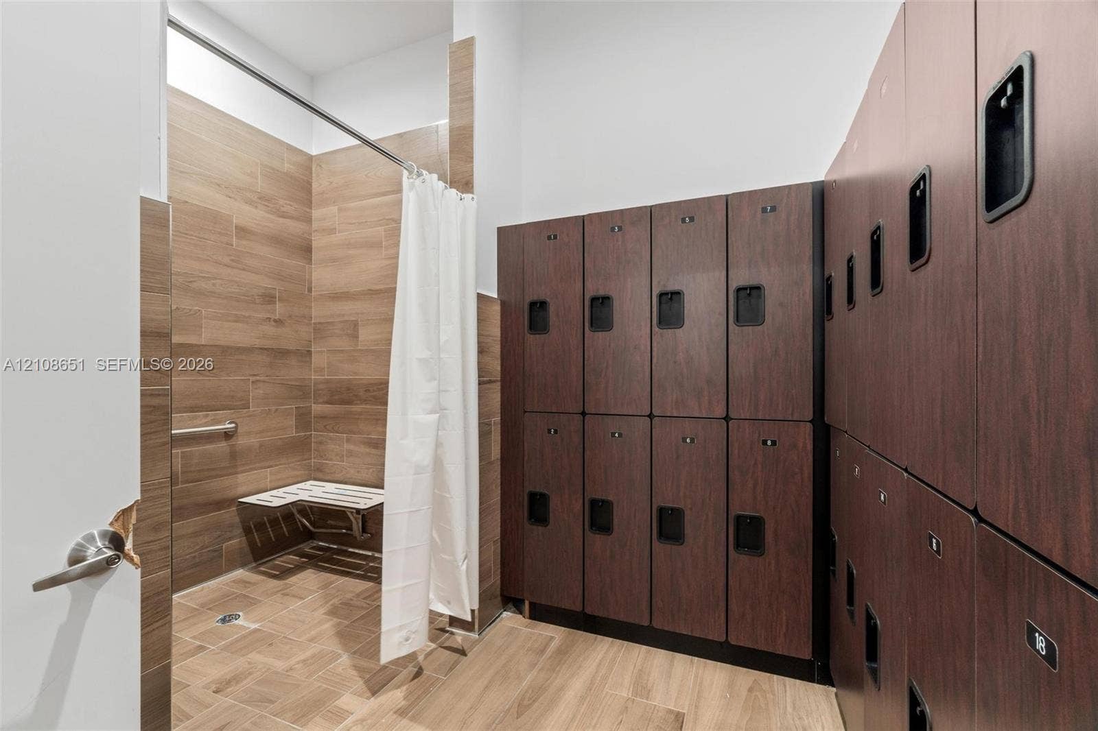 3288 W 106th Terrace, #3288, Hialeah, Hialeah, FL 33018 Walk-in shower featuring wood-finish tile wall and floor surfaces, wall-mounted bench, and integrated metal grab bar, accompanied by a series of built-in dark wood-finish storage lockers