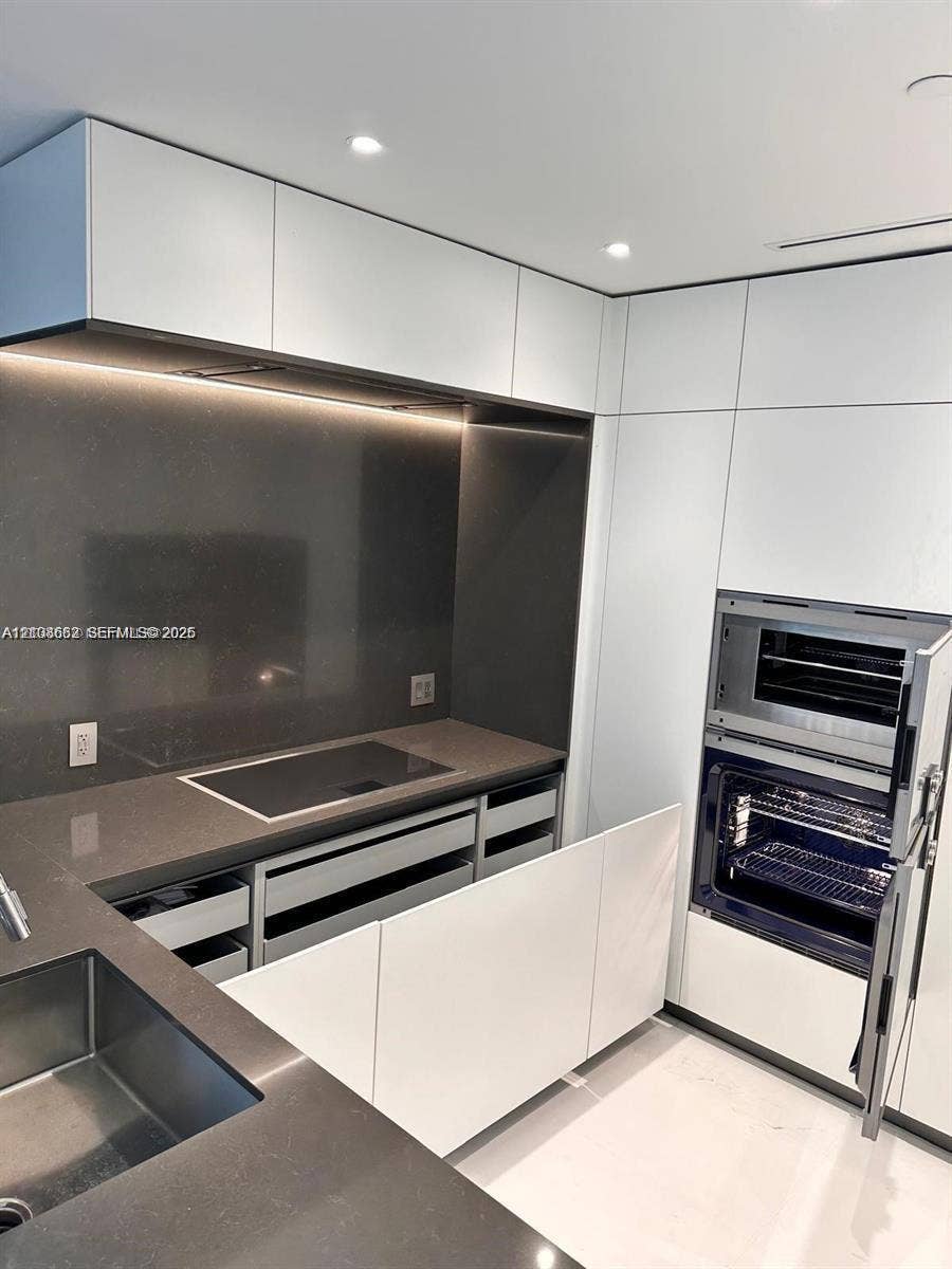 300 Biscayne Boulevard, #2709W, Miami, Miami, FL 33131 Modern kitchen featuring white flat-panel cabinetry, dark stone countertops, integrated induction cooktop, built-in dual oven, and undermount stainless steel sink