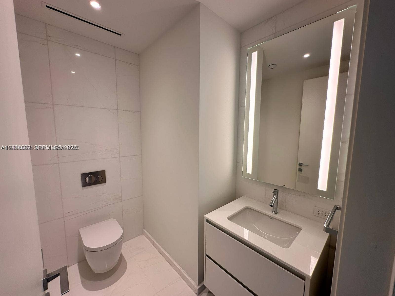 300 Biscayne Boulevard, #2709W, Miami, Miami, FL 33131 Wall-mounted toilet with a flush plate, vanity featuring a rectangular basin, integrated mirror with vertical LED lighting, and large-format porcelain tile wall cladding