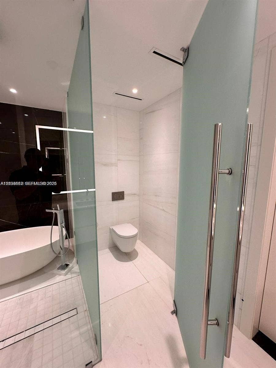 300 Biscayne Boulevard, #2709W, Miami, Miami, FL 33131 Freestanding soaking tub with floor-mounted chrome faucet, wall-hung toilet, large-format tile flooring, and frosted glass partition with polished chrome hardware