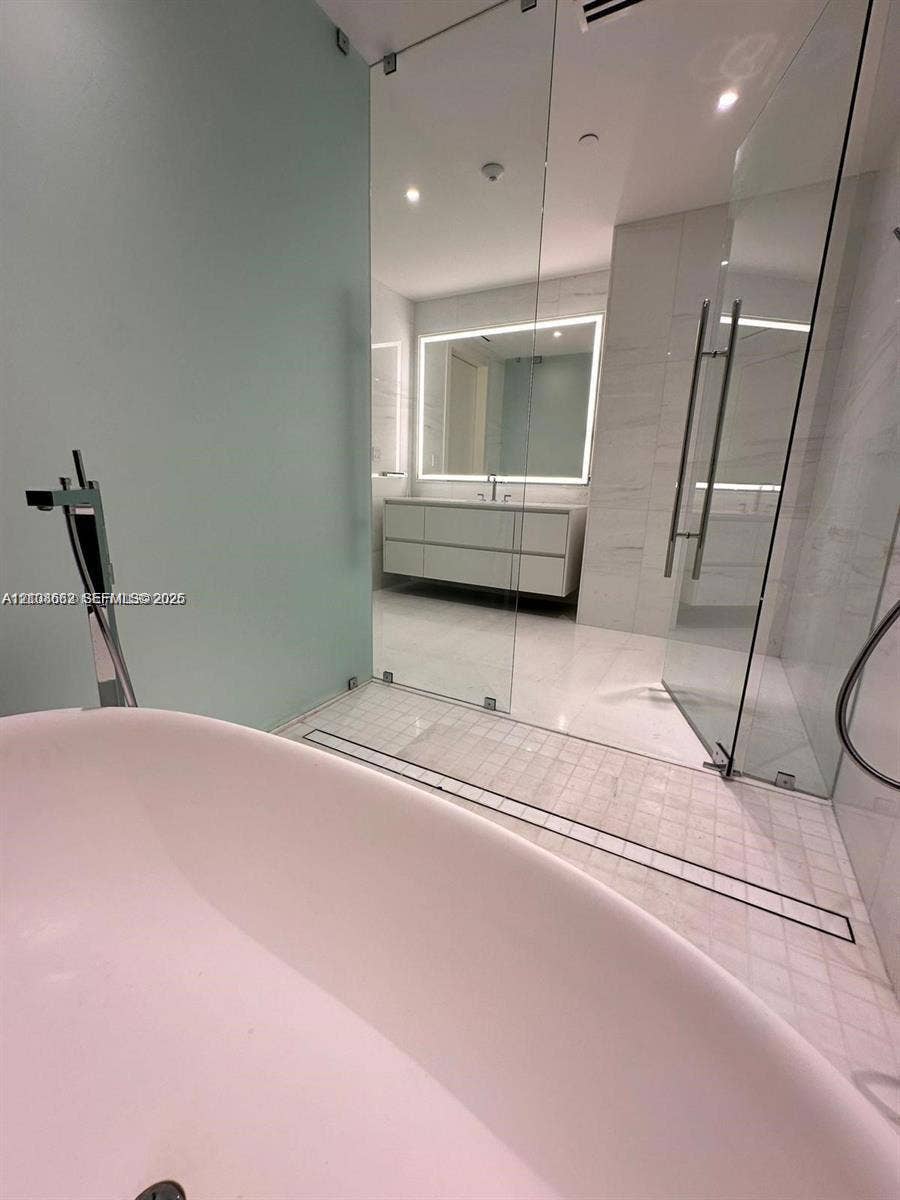 300 Biscayne Boulevard, #2709W, Miami, Miami, FL 33131 Freestanding soaking tub with a floor-mounted faucet, frameless glass-enclosed shower, mosaic tile shower flooring, wall-mounted double vanity with a backlit mirror, and large-format tile flooring