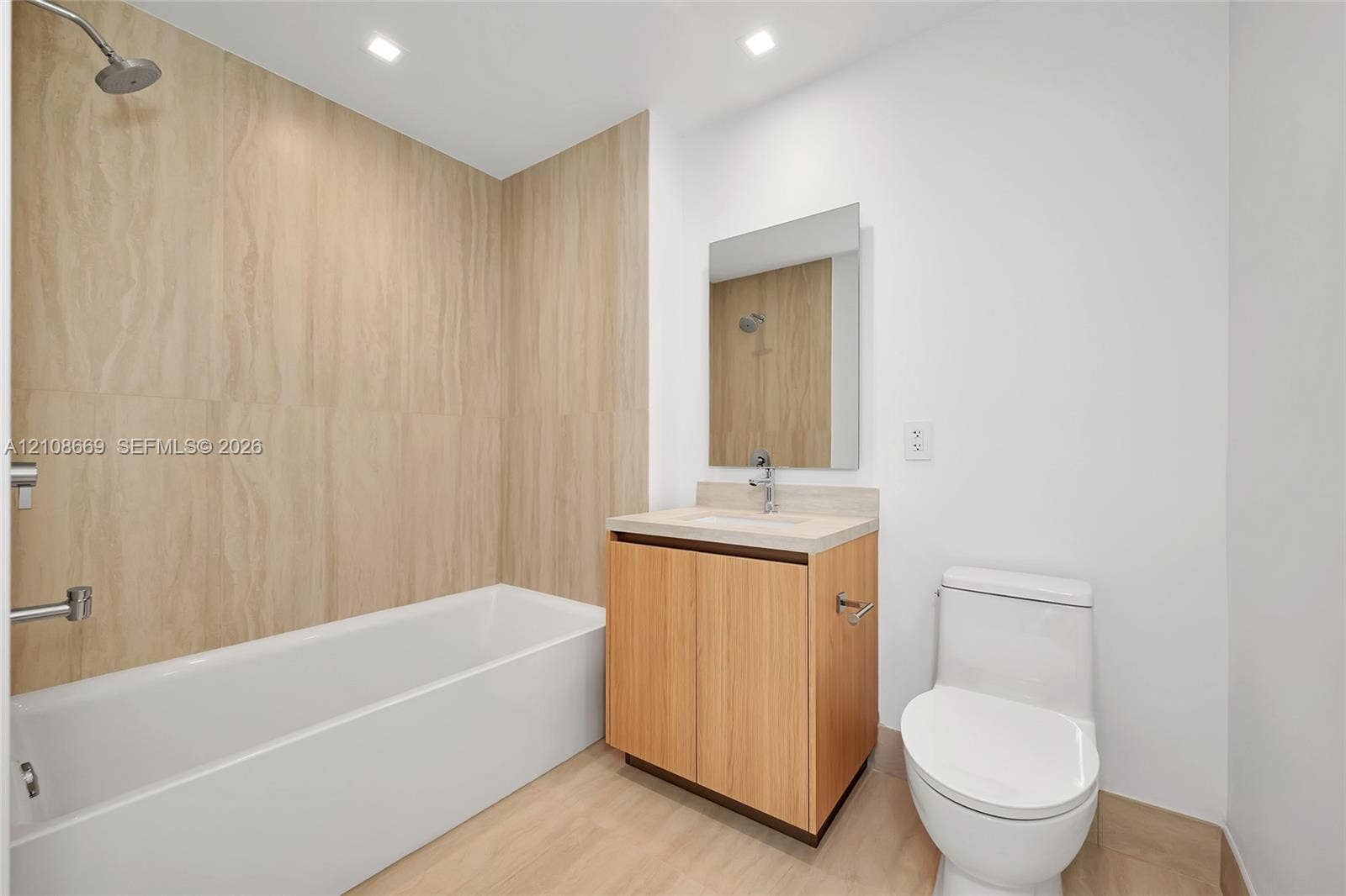 Bathroom featuring a wood-finish vanity with a stone countertop, a white soaking tub with floor-to-ceiling textured tile surround, a wall-mounted mirror, and recessed lighting