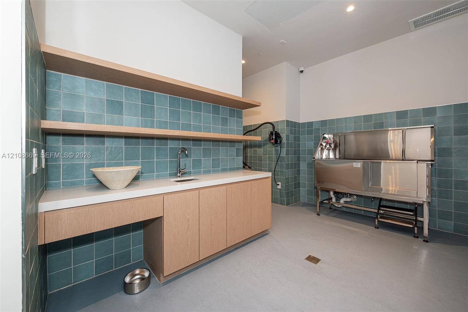 Custom cabinetry with light wood-finish detailing, quartz countertop, square-tiled wall backsplash, open shelving, and stainless steel basin