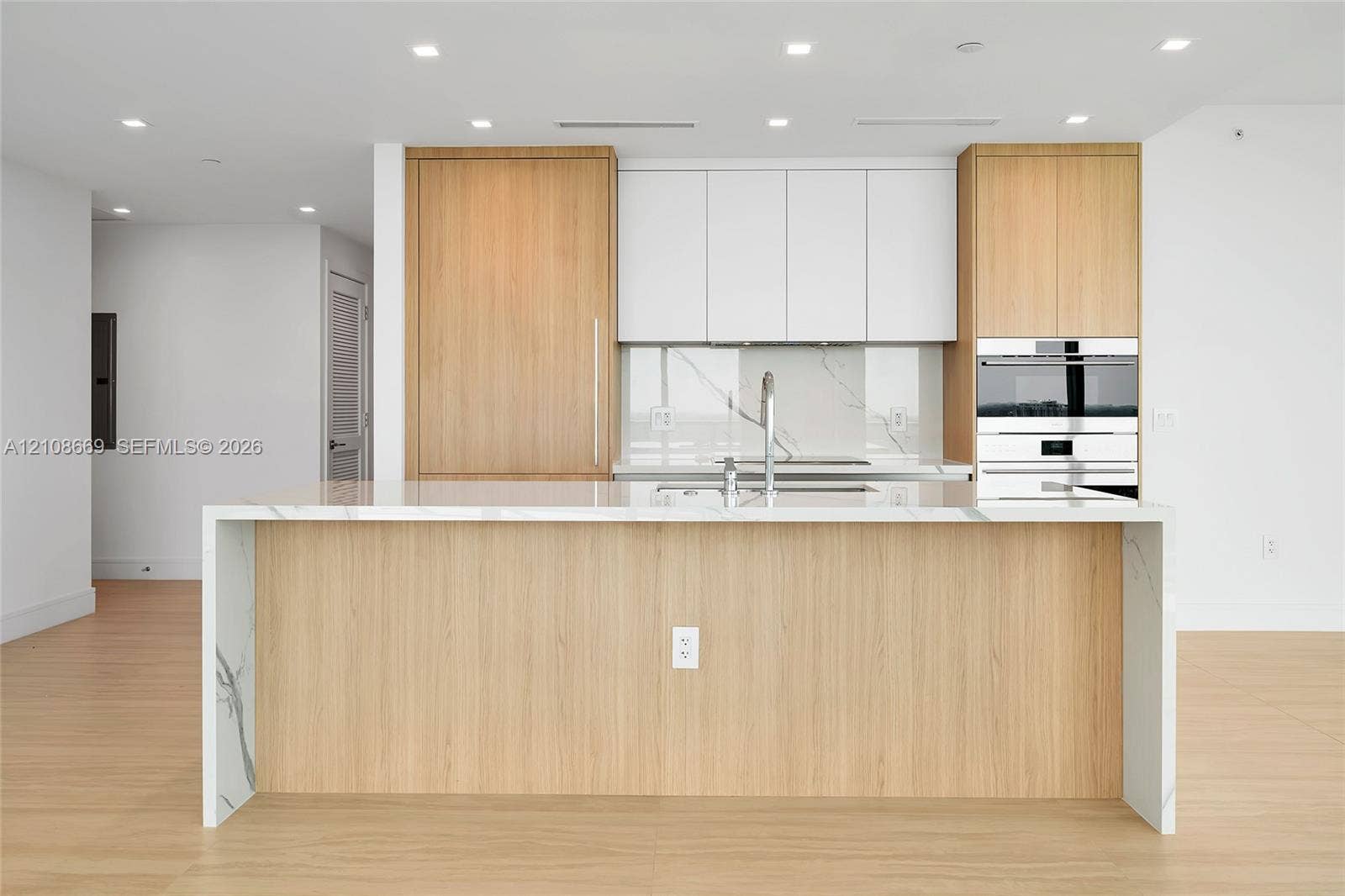 Kitchen featuring a waterfall-edge island, wood-finish cabinetry, white upper cabinets, stone-surfaced countertops and backsplash, and built-in stainless steel appliances