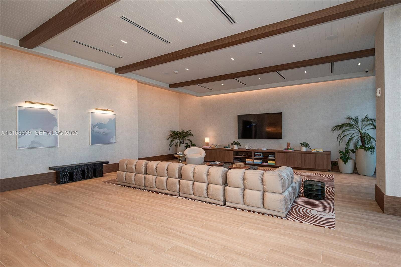Large living area featuring wood-finish flooring, recessed ceiling lighting, decorative wood-beam ceiling accents, wall-integrated cove lighting, and custom built-in cabinetry