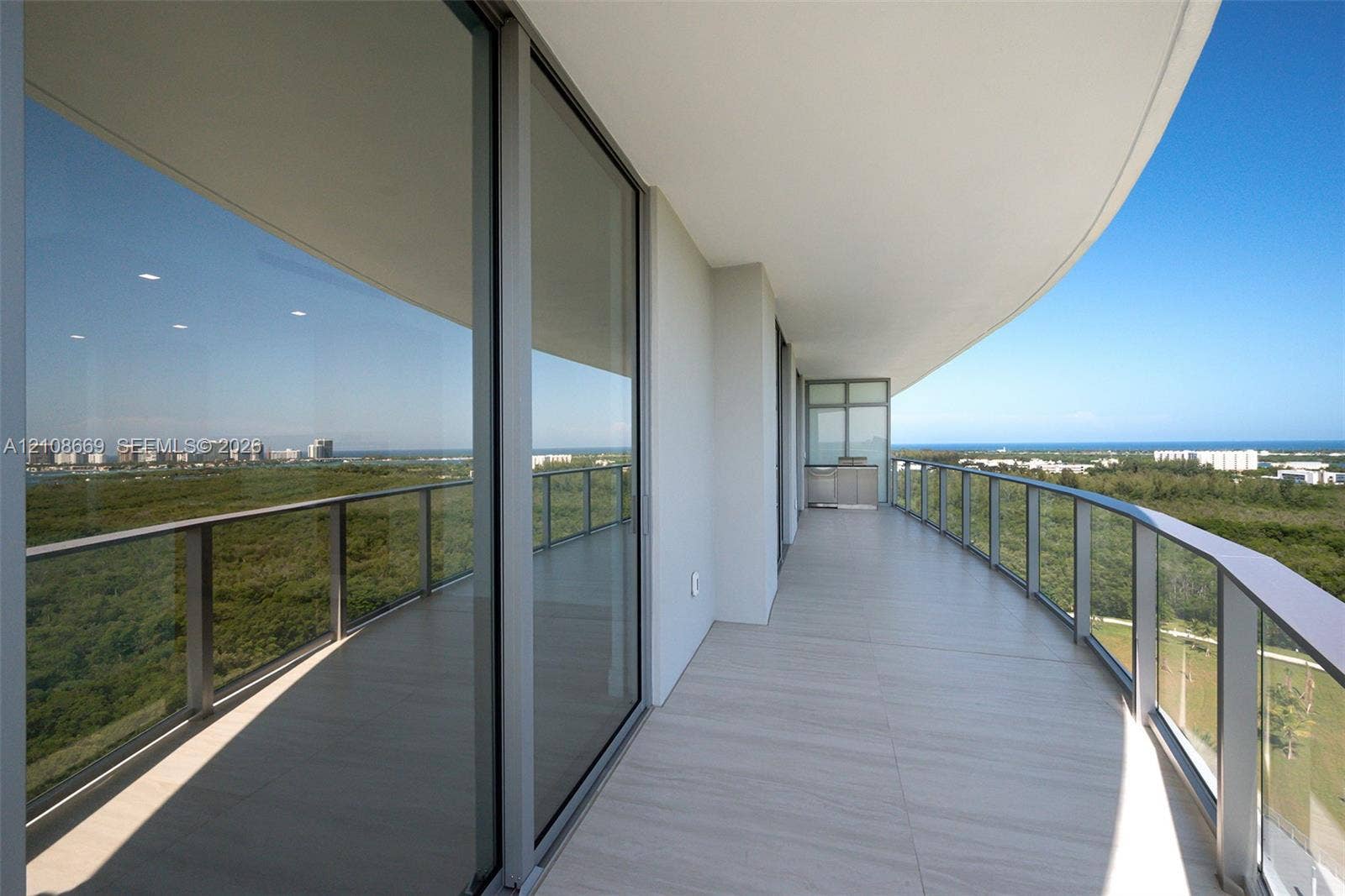 Curved covered balcony featuring large-format tile flooring, a glass-panel railing, floor-to-ceiling sliding glass doors, and a built-in outdoor kitchen station