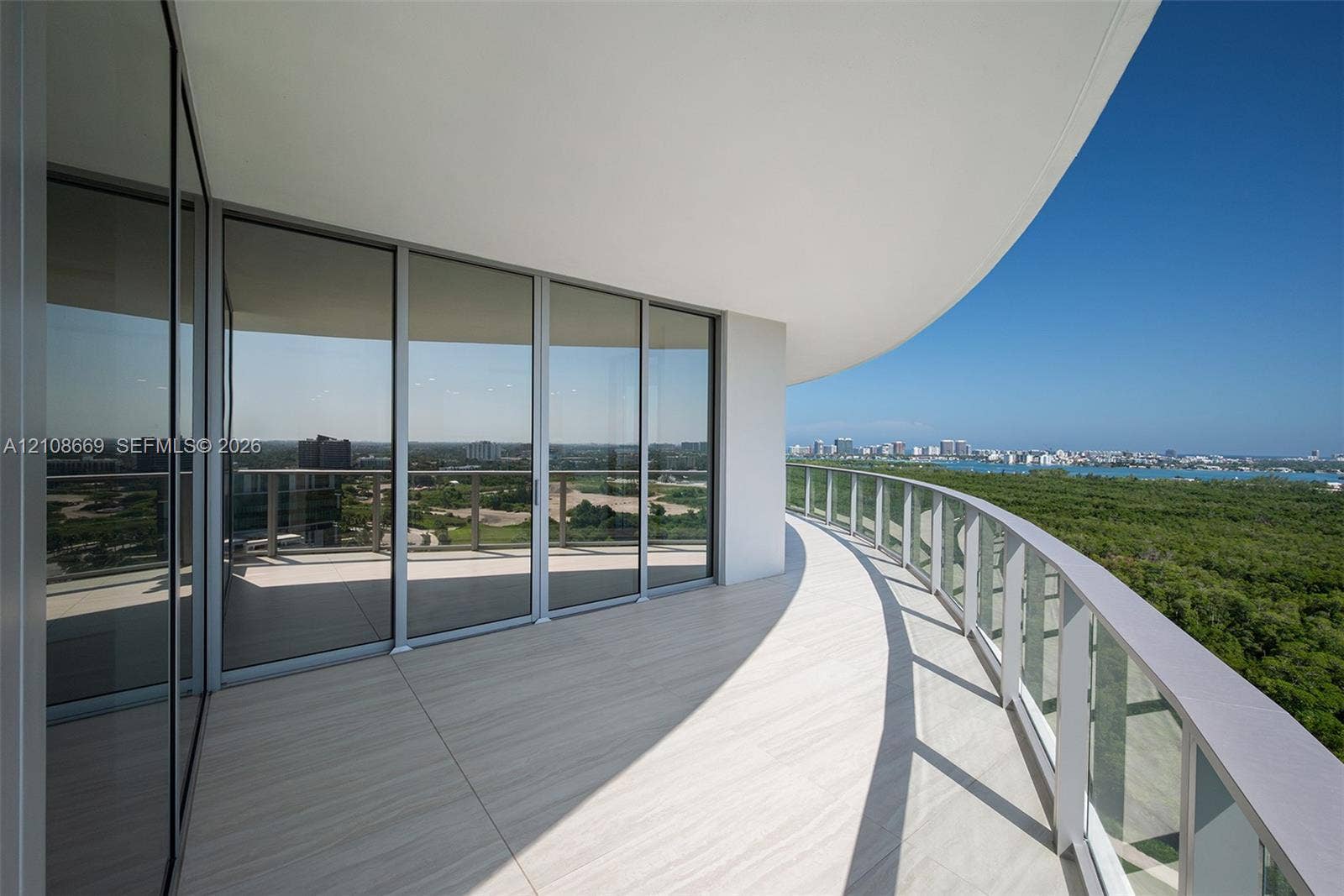 Curved balcony featuring light-toned tile flooring, glass panel railing, floor-to-ceiling sliding glass doors, and a deep overhang