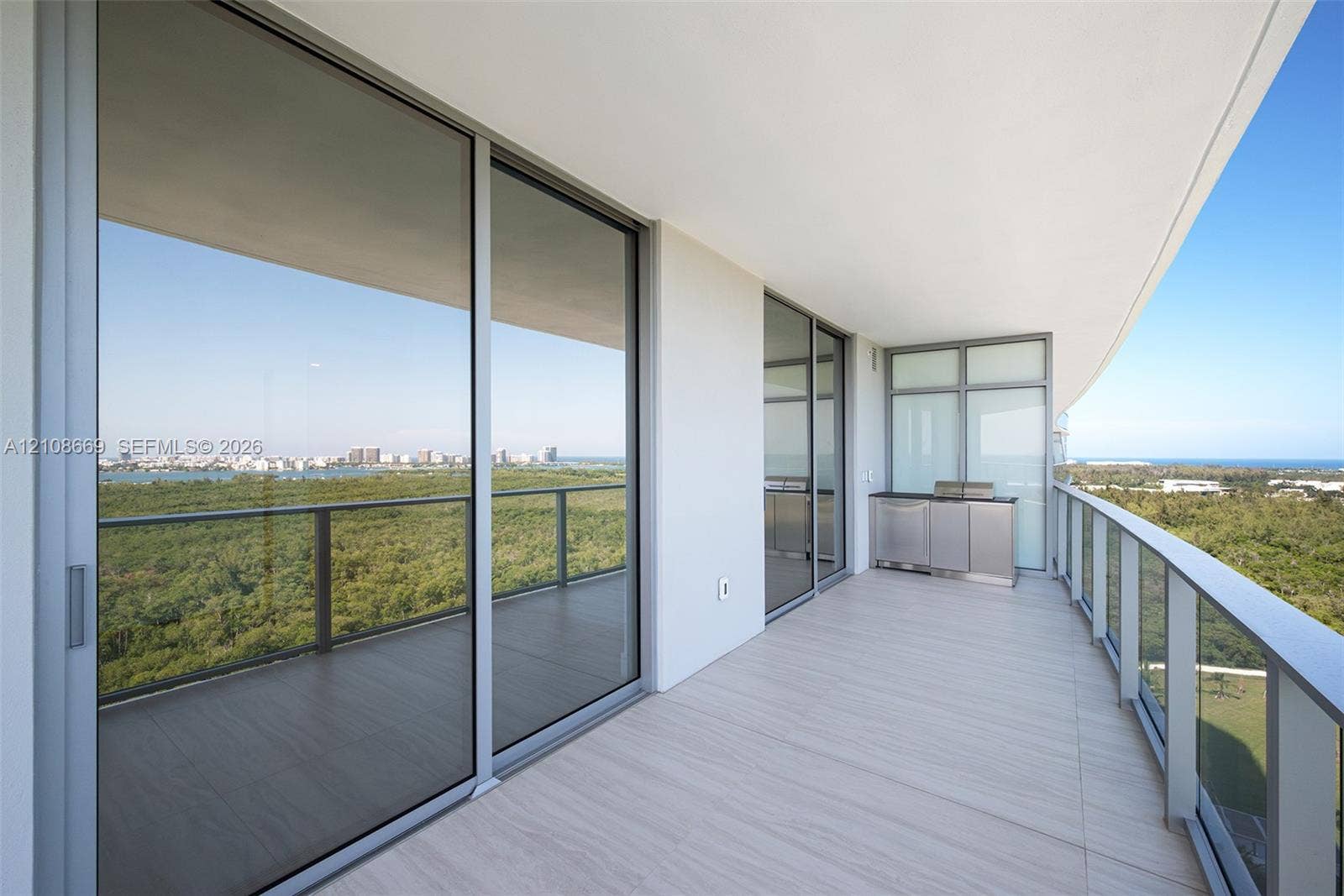 Expansive wrap-around balcony featuring porcelain tile flooring, stainless steel outdoor kitchen cabinetry, glass panel railing, and floor-to-ceiling sliding glass doors