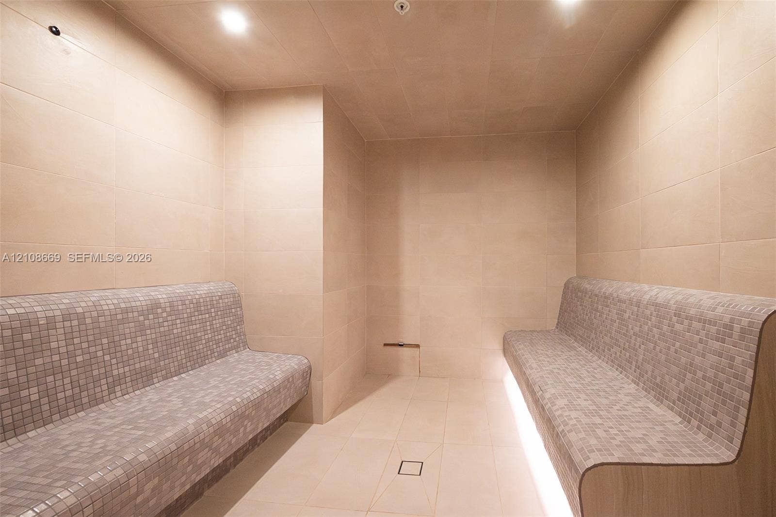 Steam room featuring large-format wall and floor tiling, dual built-in benches with mosaic tile surfacing, and recessed toe-kick lighting