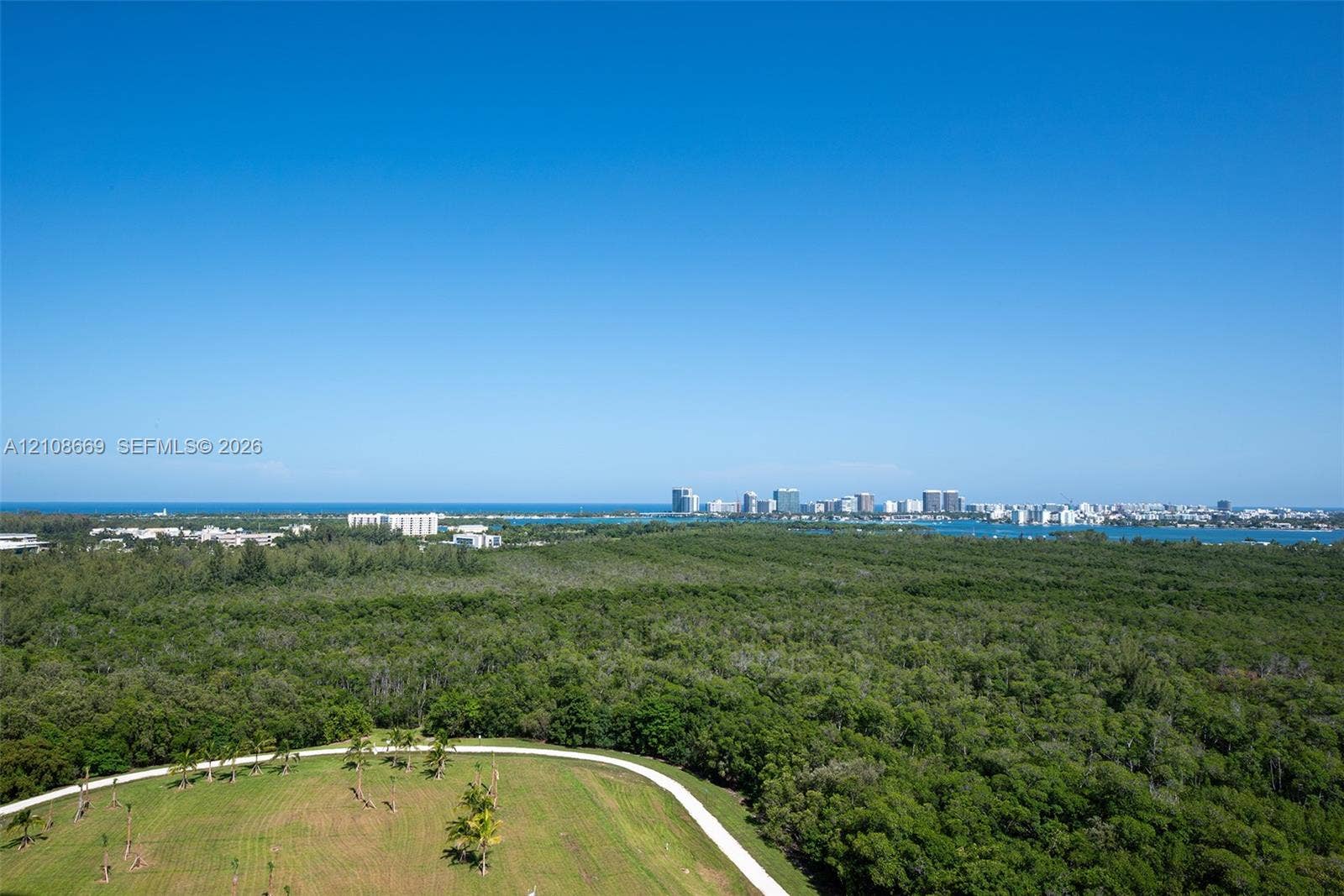 Aerial perspective showcasing extensive natural greenery, open lawn space, paved pathways, coastal water frontage, and a distant cityscape