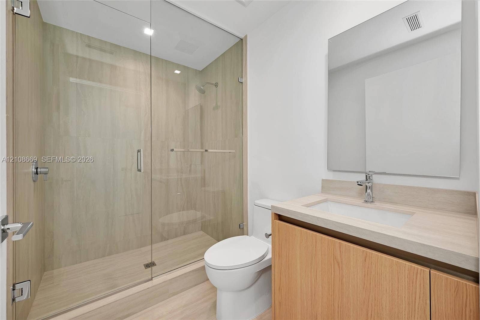 Full-height glass shower enclosure with integrated light-toned tile, wood-finish cabinetry, countertop-integrated basin, and frameless wall mirror