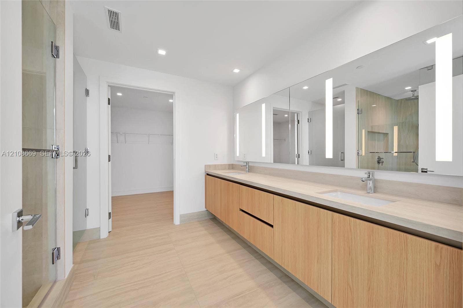 Dual-sink vanity with wood-finish cabinetry, countertop with integrated rectangular basins, large wall-mounted mirrors with integrated vertical lighting, and a glass-enclosed shower