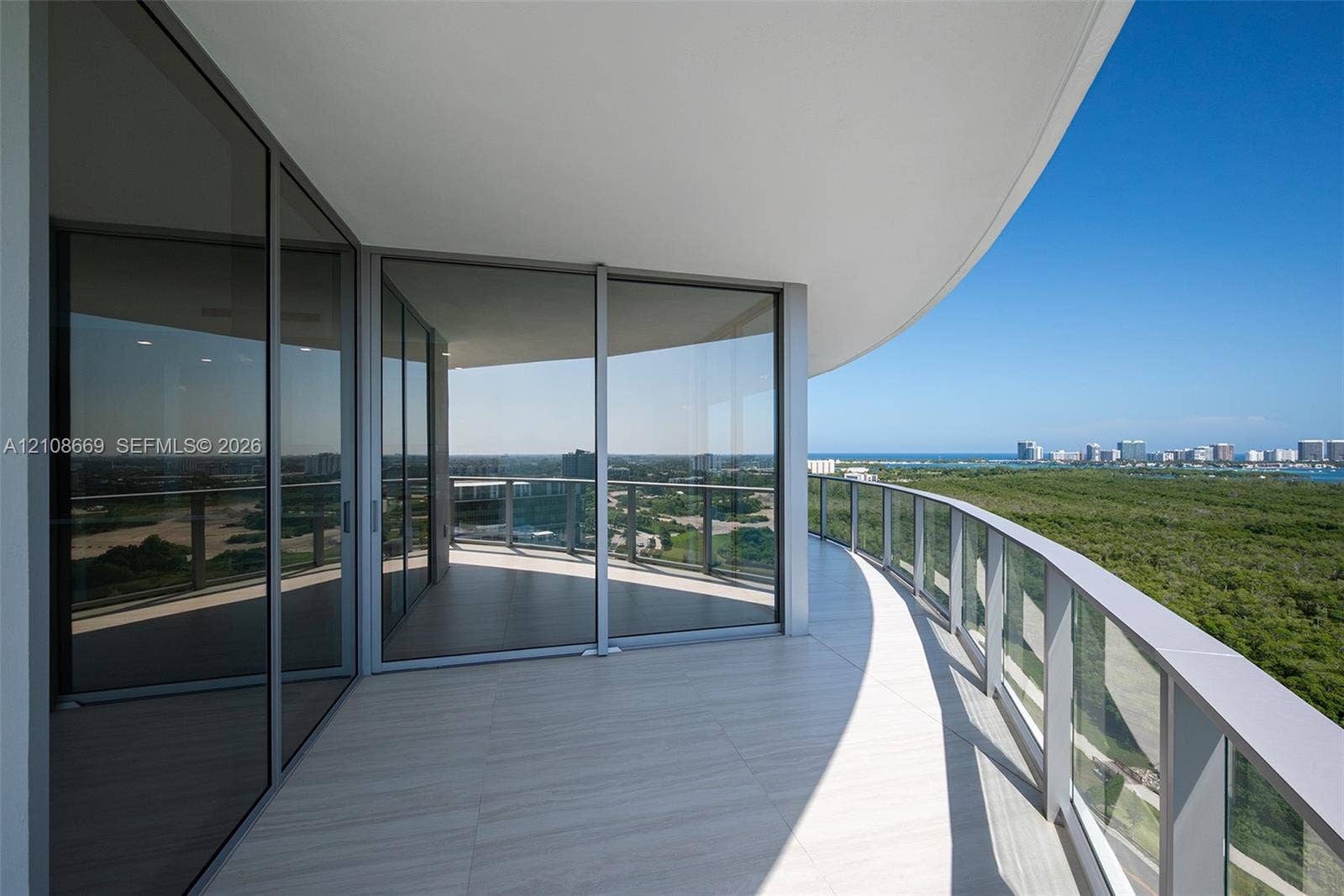 Expansive curved balcony featuring floor-to-ceiling glass doors, neutral tile flooring, and a sleek glass-panel railing
