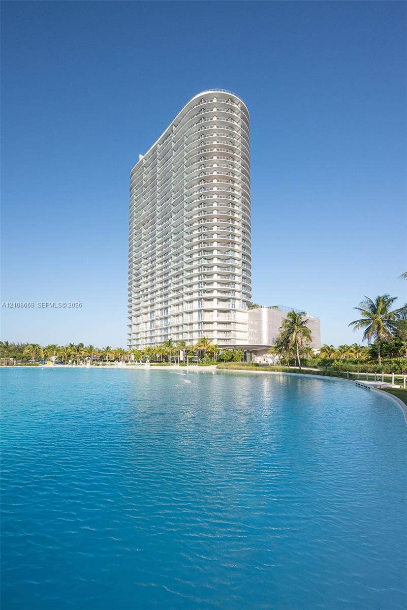 High-rise tower featuring rounded architectural design and expansive exterior balconies, situated along a large man-made waterfront with tropical landscaping