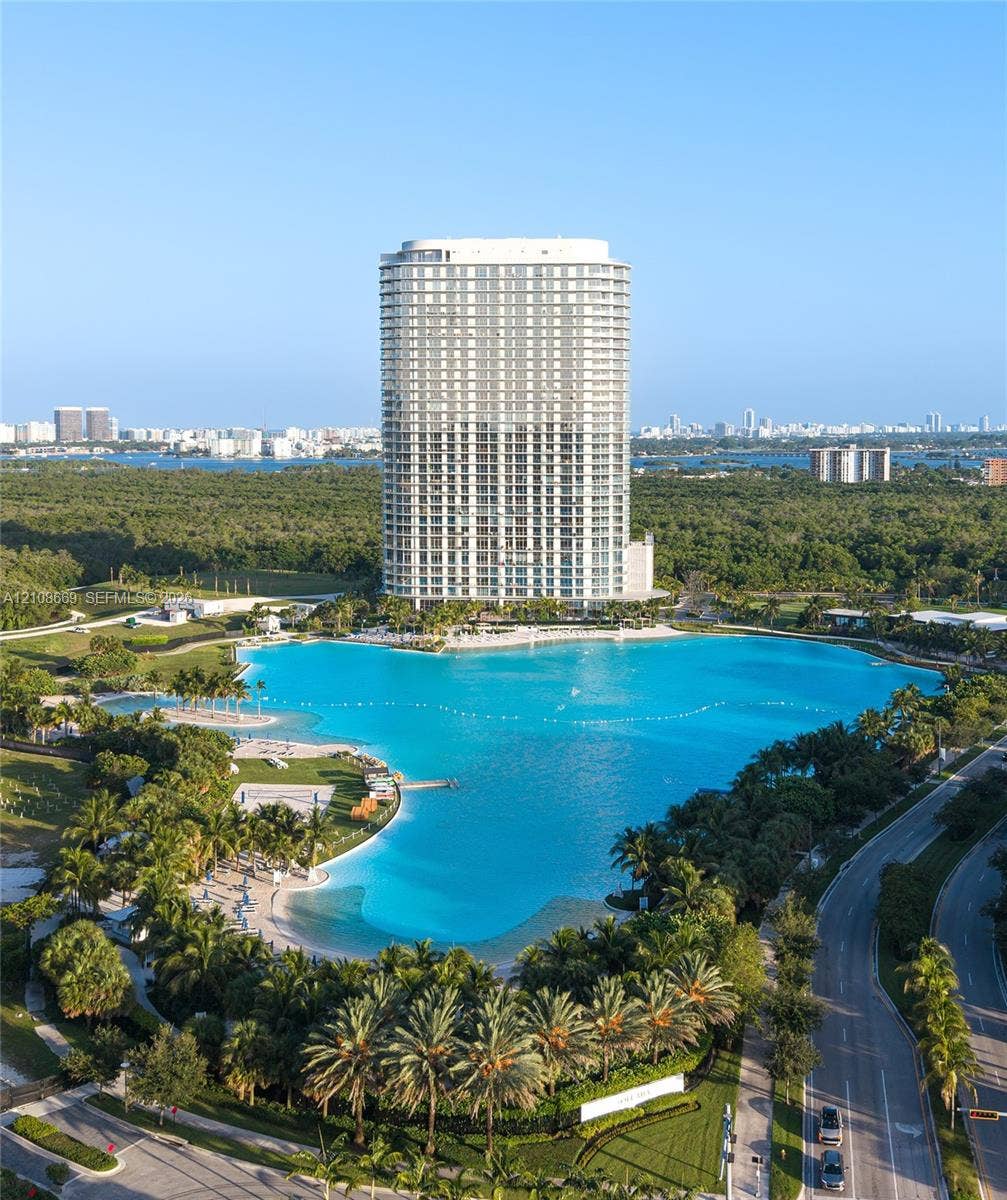 High-rise residential tower featuring floor-to-ceiling glass windows, a large man-made lagoon with sandy shorelines, and landscaped grounds with mature palm trees