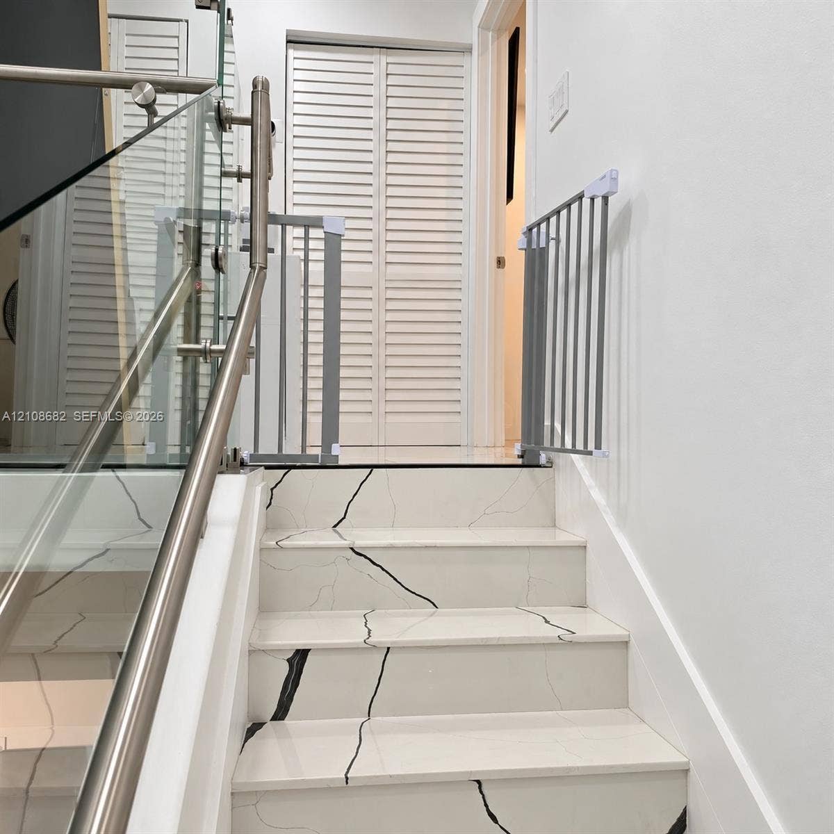 8943 NW 112th Terrace, Hialeah Gardens, FL 33018 Staircase featuring white stone-finish treads with dark veining, stainless steel handrails, and a glass balustrade, complemented by white louvered closet doors