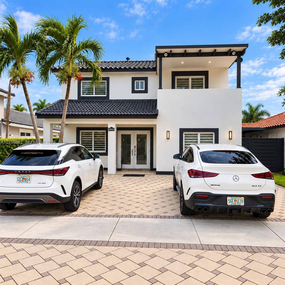 8943 NW 112th Terrace, Hialeah Gardens, FL 33018 Two-story exterior featuring a high-contrast stucco facade, dark roof tiles, a covered entry with French doors, a second-level balcony, and an expansive paved driveway