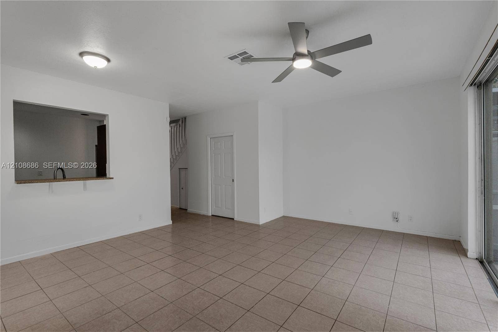 Neutral tile flooring, ceiling fan with integrated lighting, pass-through opening to kitchen, and sliding glass door