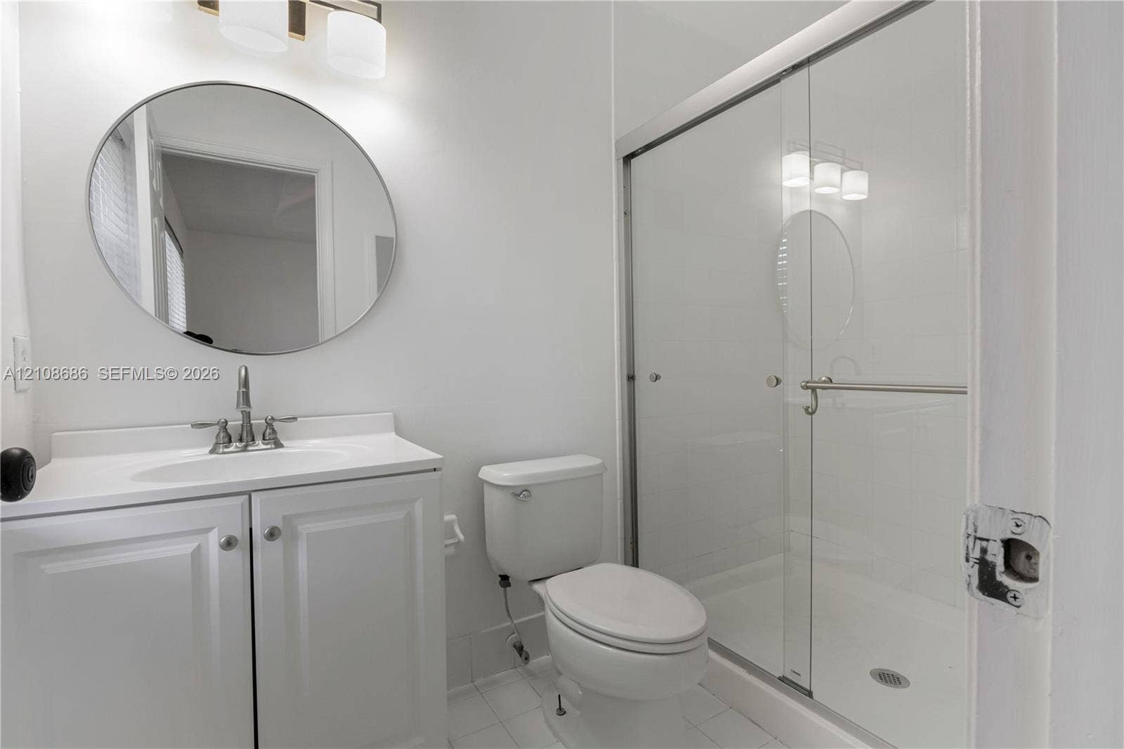 Bathroom featuring a white vanity cabinet, circular wall-mounted mirror, three-light fixture, and a glass-enclosed shower