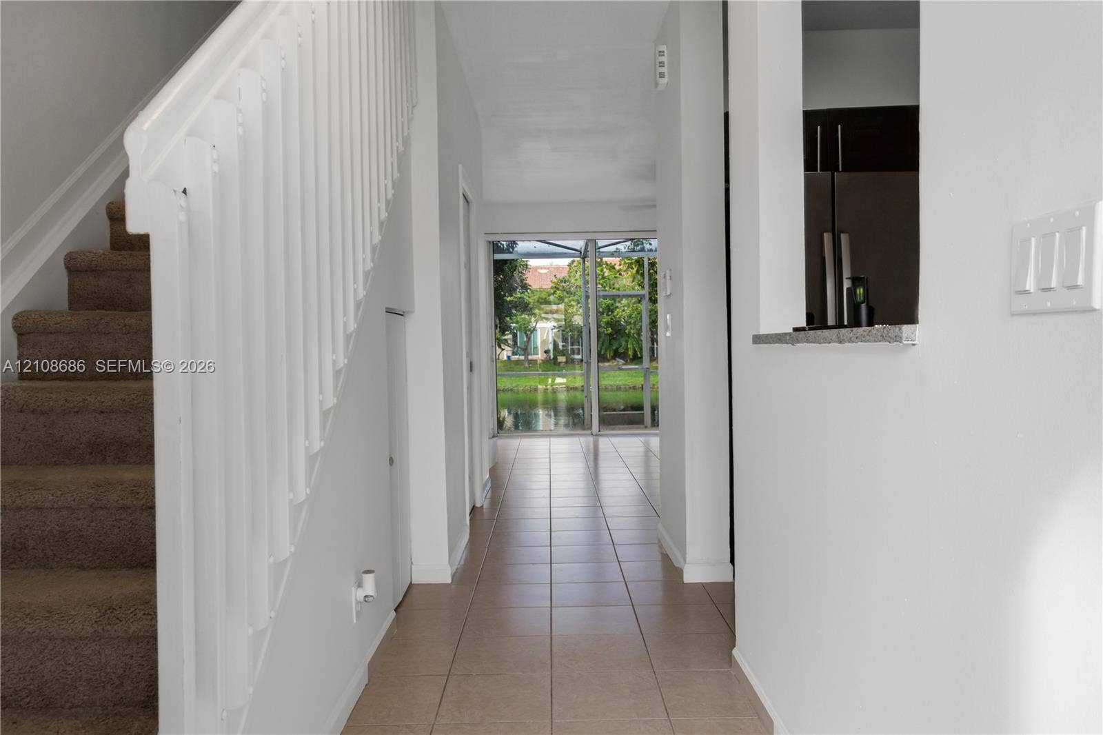 Interior hallway featuring ceramic tile flooring, a staircase with a white balustrade, and a pass-through opening to the kitchen area