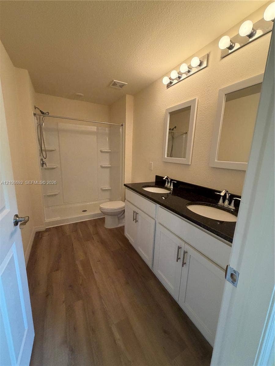 Dual-sink vanity with dark stone-finish countertop, white cabinetry, wood-finish flooring, and white shower-tub surround