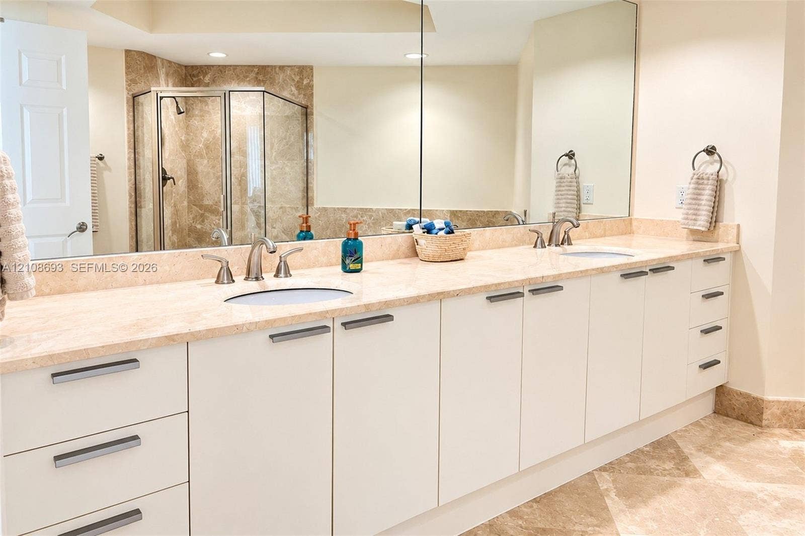 231 Riverside Drive, #1210, Daytona Beach, Daytona Beach, FL 32117 Extended dual-sink vanity featuring white flat-panel cabinetry, stone countertops, integrated sink basins, and polished metal hardware, accompanied by a large wall-mounted mirror, recessed lighting, and a walk-in shower with natural stone tile surrounds