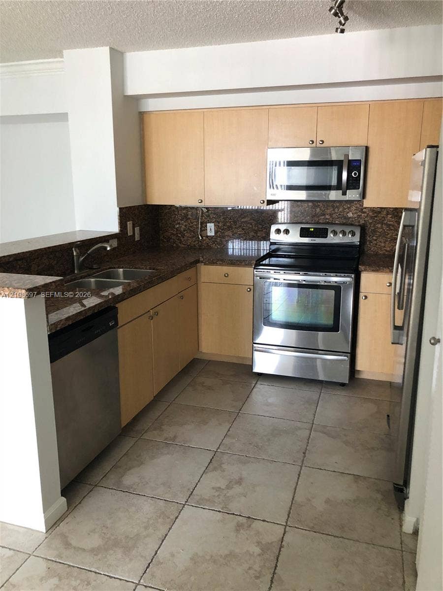 Kitchen featuring light wood-finish cabinetry, stone-finish countertops, stainless steel appliances, a double-basin sink, and tile flooring