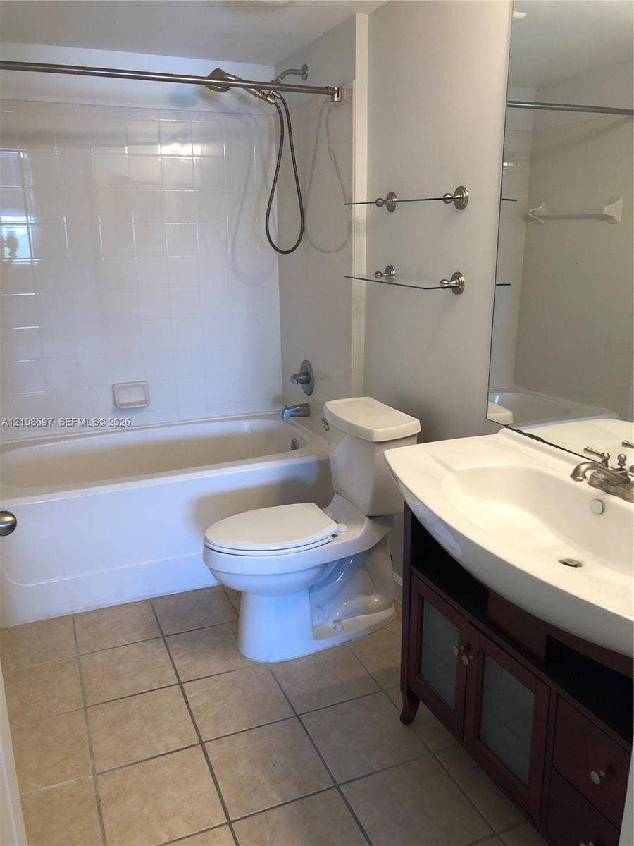 Bathroom featuring a white bathtub with a tile surround, vanity with an integrated sink, glass shelving, and tile flooring
