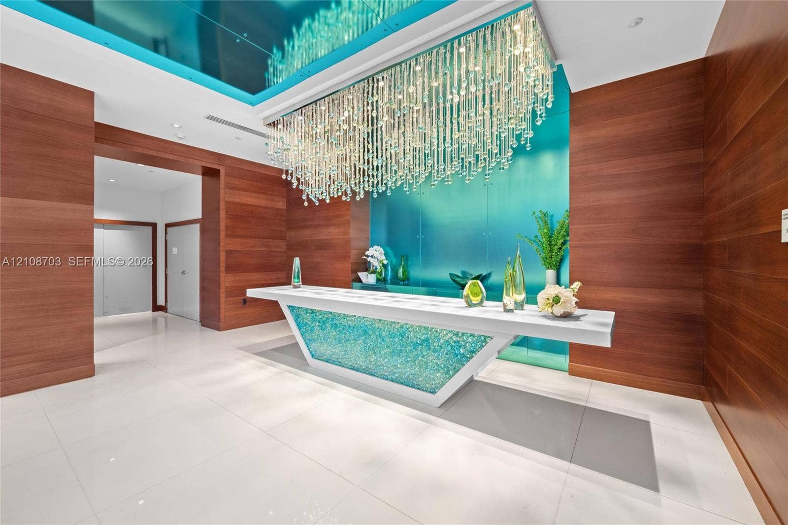 300 Sunny Isles Boulevard, #4-1501, Sunny Isles Beach, Sunny Isles Beach, FL 33160 Lobby featuring wood-paneled walls, large-format tile flooring, a geometric reception desk with illuminated glass inlay, and a multi-strand crystal chandelier