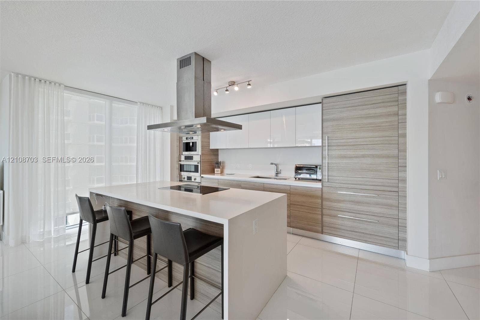 300 Sunny Isles Boulevard, #4-1501, Sunny Isles Beach, Sunny Isles Beach, FL 33160 Modern kitchen featuring an island with a waterfall-edge countertop, floor-to-ceiling cabinetry with wood-grain finish, integrated appliances, a stainless steel chimney-style range hood, and large-format tile flooring