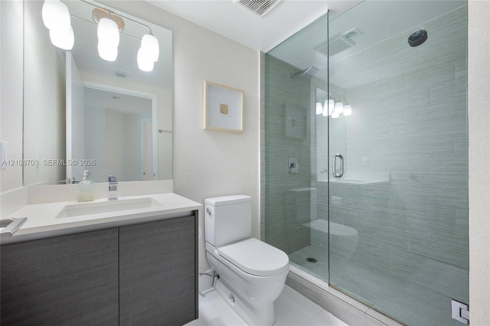 300 Sunny Isles Boulevard, #4-1501, Sunny Isles Beach, Sunny Isles Beach, FL 33160 Bathroom featuring a dark wood-finish vanity with a light-toned countertop, a frameless glass-enclosed walk-in shower with textured tile surround, and a wall-mounted light fixture above a large mirror