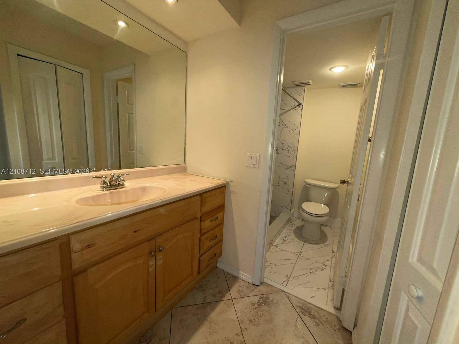 379 Lakeview Drive, #205, Weston, Weston, FL 33326 Bathroom featuring wood-finish cabinetry, an expansive wall-mounted vanity mirror, ceramic tile flooring, and a separate water closet area with patterned tile surrounds