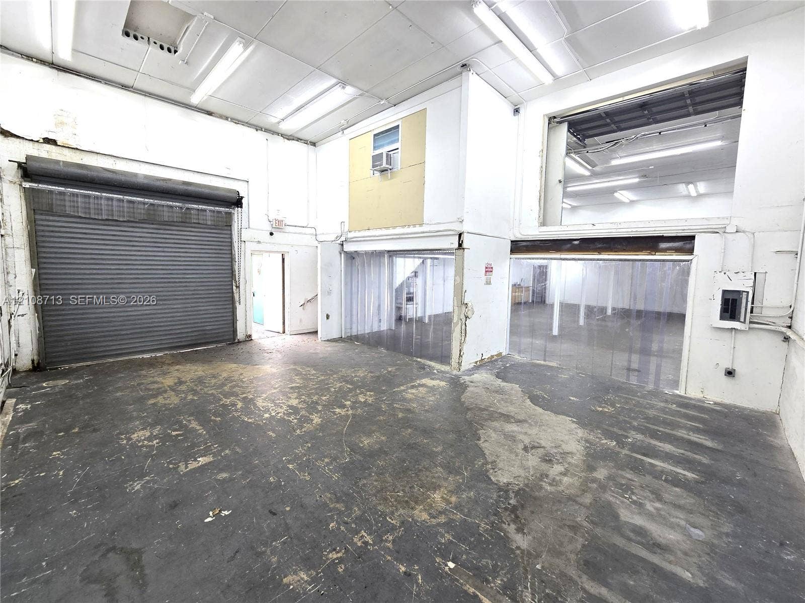 Industrial workspace featuring a high-clearance metal roll-up door, multi-level layout, exposed ceiling-mounted fluorescent lighting, and concrete flooring