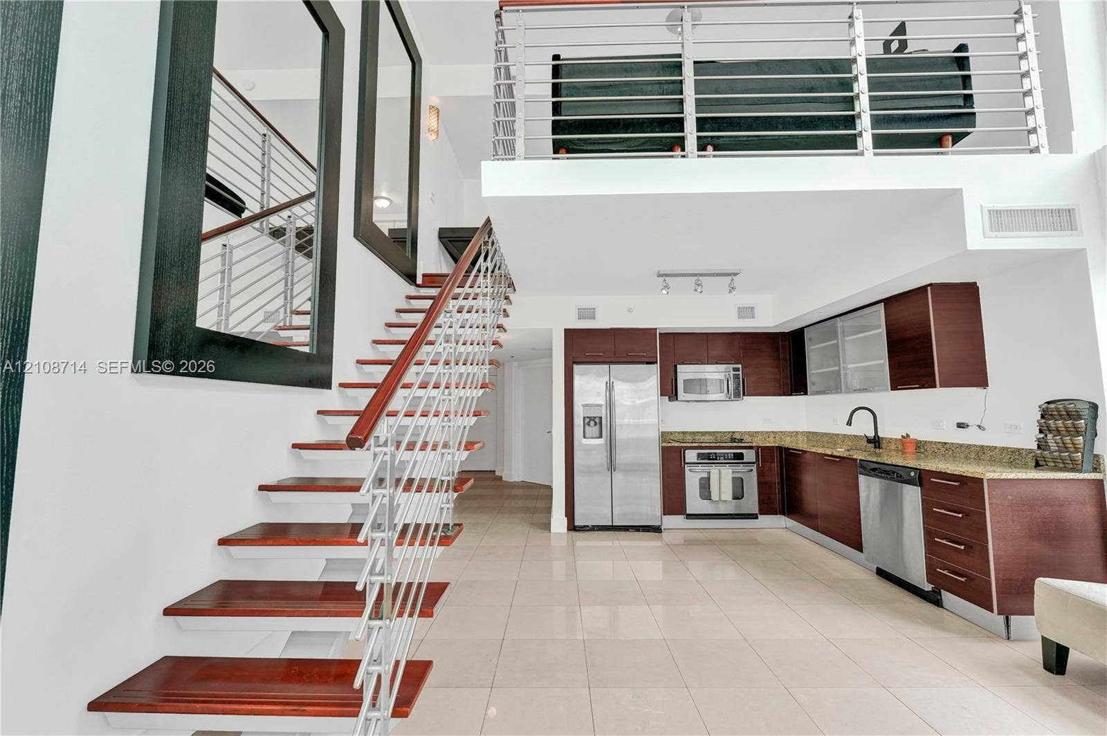 41 SE 5th Street, #317, Miami, Miami, FL 33131 Open-concept interior featuring a stainless steel appliance suite, rich-toned cabinetry with stone-finish countertops, ceramic tile flooring, a mezzanine level with metal railings, and a floating staircase with wood-finish treads and metal cabling