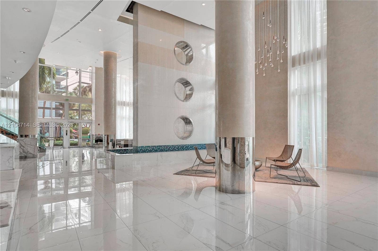 41 SE 5th Street, #317, Miami, Miami, FL 33131 Grand lobby featuring expansive tile flooring, double-height ceilings, structural support columns with metallic accents, a decorative vertical wall feature with circular insets, and a suspended multi-light chandelier
