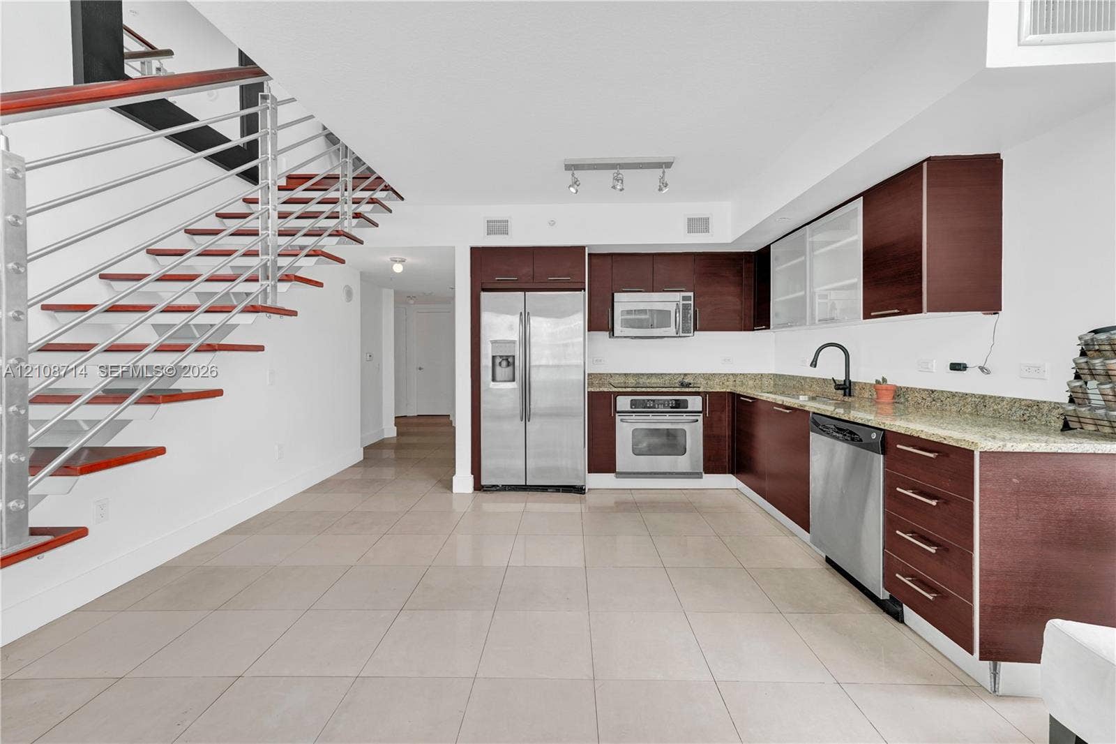 41 SE 5th Street, #317, Miami, Miami, FL 33131 Kitchen featuring wood-finish cabinetry, stone countertops, stainless steel appliances, and large-format tile flooring, accented by an open-riser staircase with a horizontal cable railing system