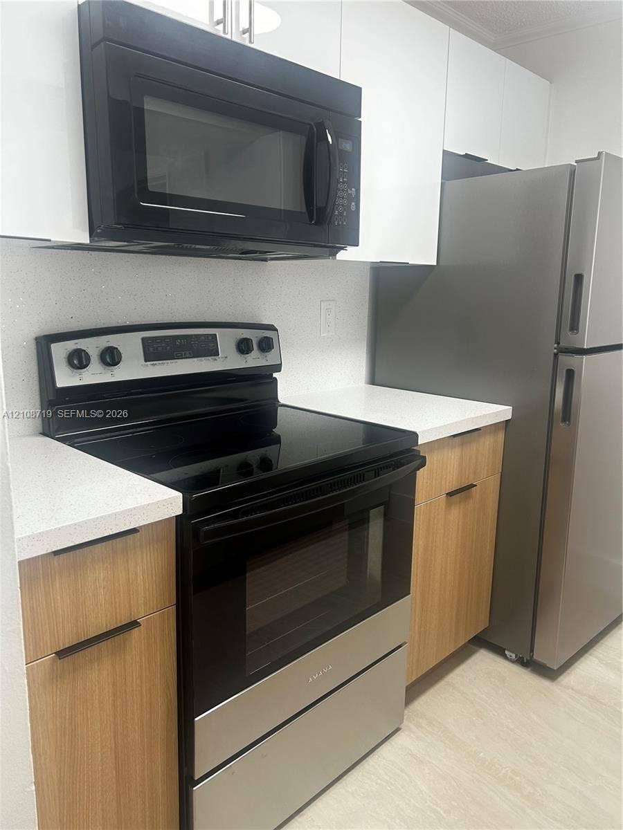 7035 NW 186th Street, #D309, Hialeah, Hialeah, FL 33015 Kitchen featuring light wood-finish cabinetry, white stone-composite countertops, stainless steel refrigerator, black electric range, and over-the-range microwave