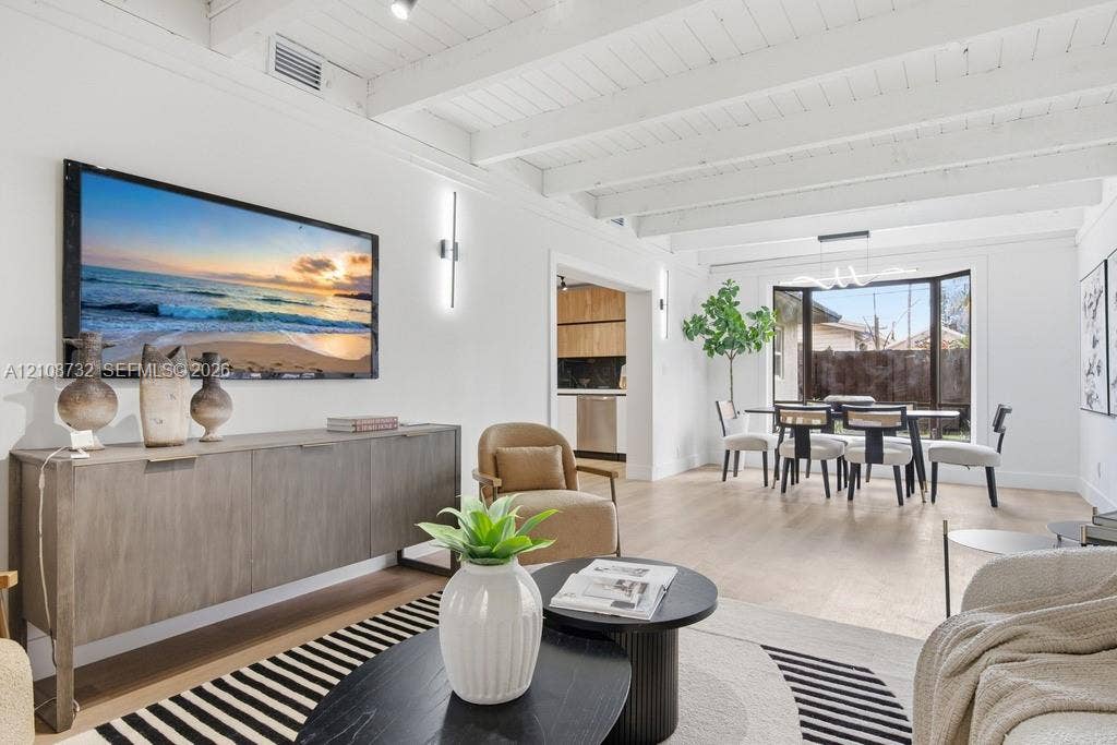 Exposed white-painted ceiling beams, wide-plank wood-finish flooring, linear wall-mounted lighting, and a bay window featuring a modern multi-light chandelier