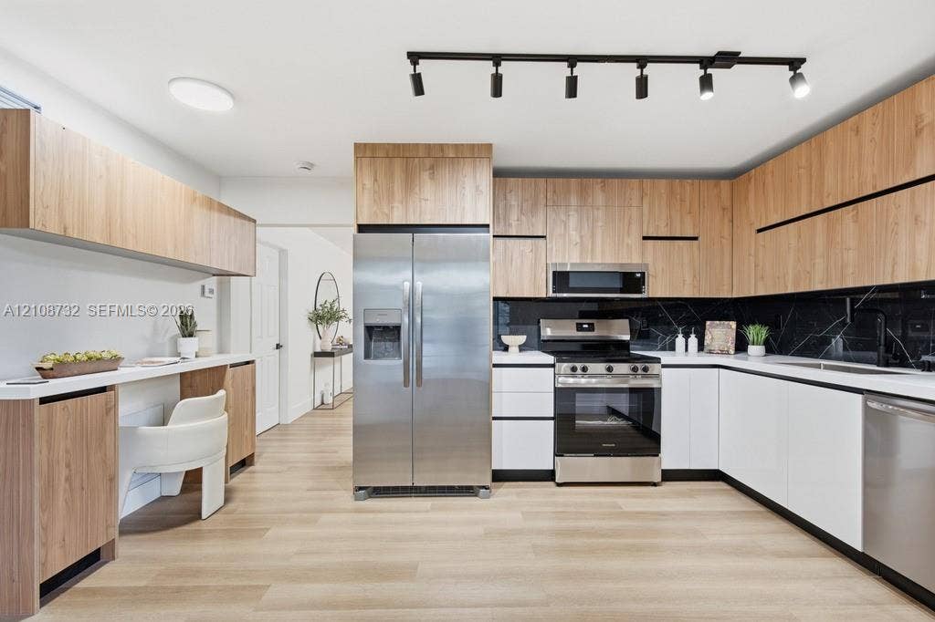Modern kitchen featuring dual-tone cabinetry, stainless steel appliances, black tile backsplash, wood-finish flooring, and industrial track lighting