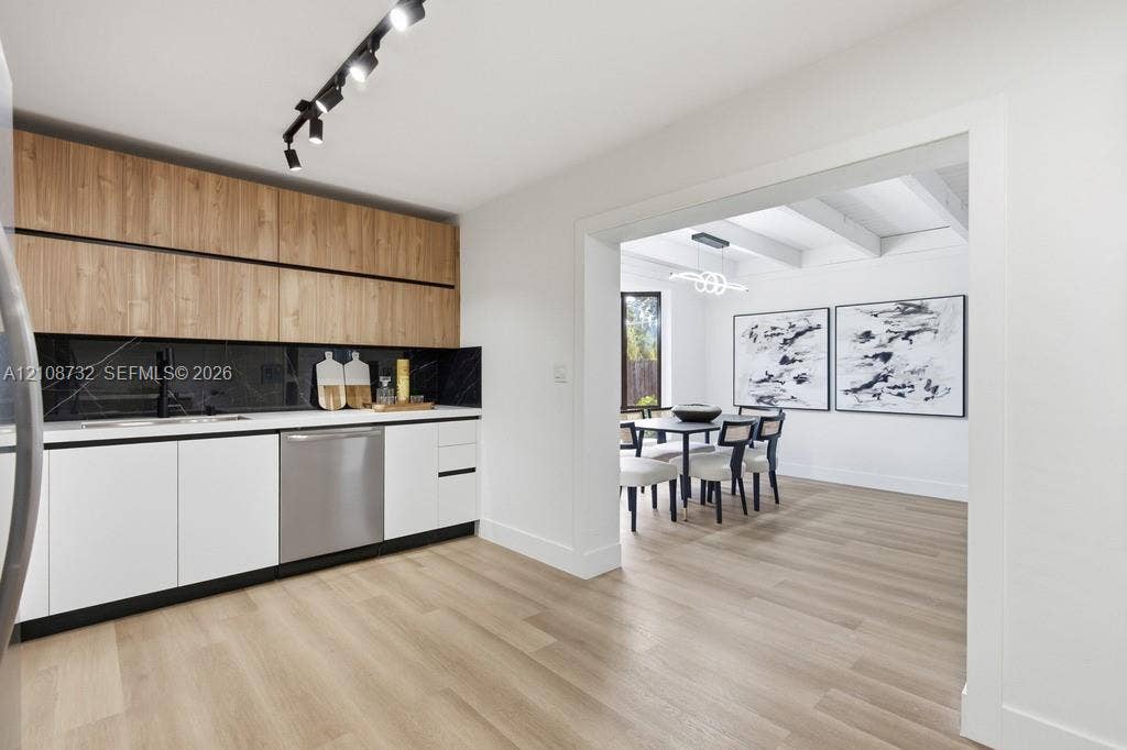 Kitchen featuring two-tone cabinetry with wood-finish upper units, dark stone-finish backsplash, stainless steel dishwasher, track lighting, and wood-finish flooring
