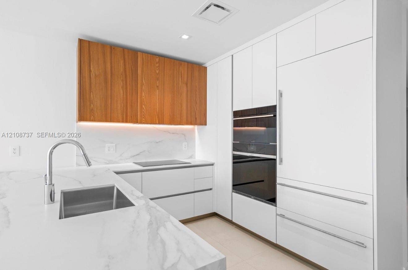 1400 Biscayne Boulevard, #3503, Miami, Miami, FL 33132 Modern kitchen featuring white flat-panel cabinetry, wood-veneer upper cabinets, light-toned stone-finish countertops and backsplash, integrated stainless steel appliances, and an undermount rectangular sink with a gooseneck faucet