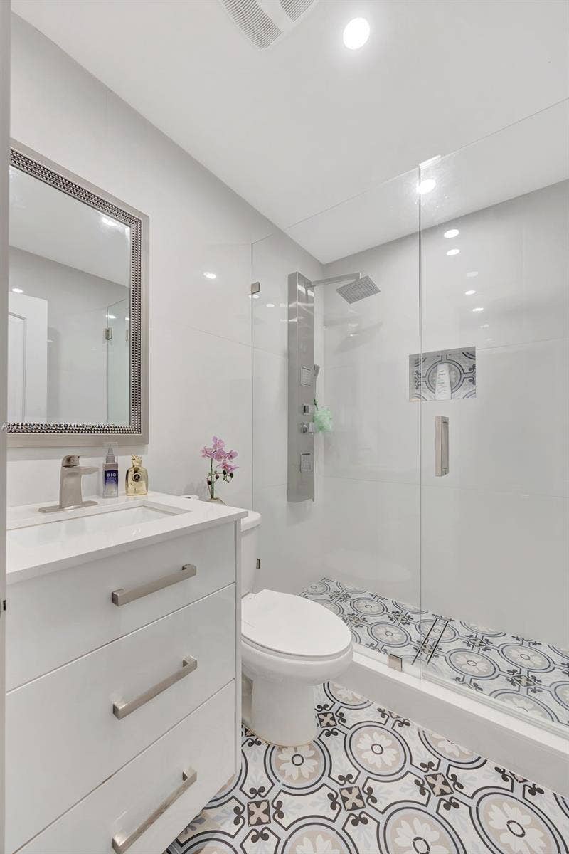 White vanity with a rectangular sink, brushed nickel hardware, patterned tile flooring, walk-in shower with a glass enclosure, and a wall-mounted shower panel