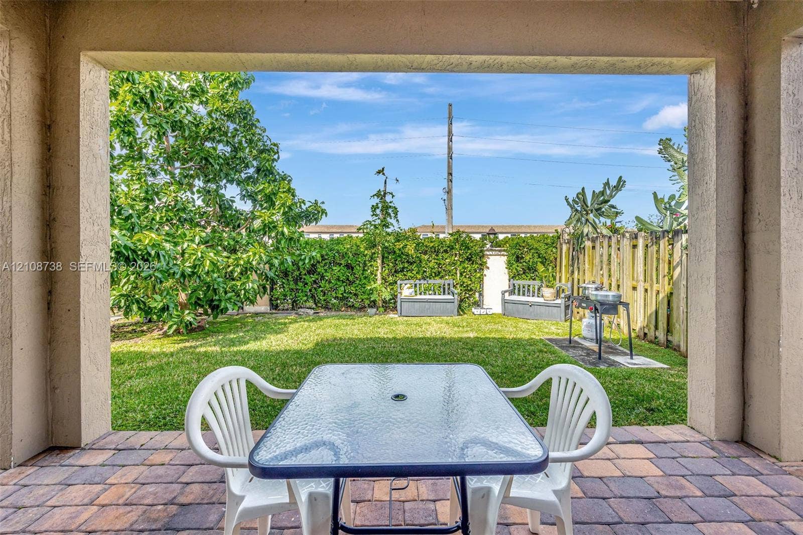 Covered patio featuring paver flooring, overlooking a backyard with a manicured lawn, mature trees, and a wooden privacy fence